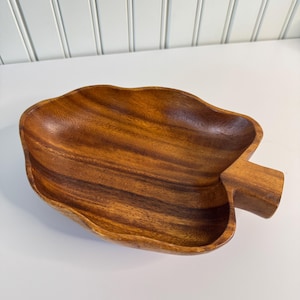 May include: A large, wooden serving bowl shaped like a leaf. The bowl is made of dark brown wood with a natural grain pattern. It has a handle on one side.