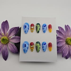 May include: Two sets of artificial nails with colourful designs are displayed on a white card, flanked by purple flowers. The nails feature blue, red, yellow, and green patterns, some with rhinestones. The nails are oval-shaped.