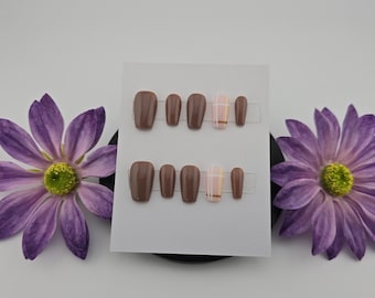 May include: Two sets of press-on nails in various shades of brown, with one accent nail featuring a pink and gold plaid pattern. The nails are displayed on a white card, flanked by two purple flowers with yellow centers.