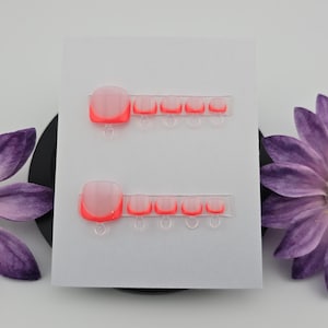 May include: Two sets of press-on toenails with a clear base and neon coral tips are displayed on a white card. The card is flanked by purple daisy-like flowers with yellow centers. The nails are a beauty product.