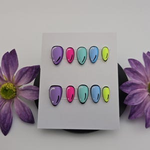 May include: A set of press-on nails in a comic book style, featuring a range of colours. Each nail is a different colour: purple, pink, teal, blue, and yellow, with black outlines. The nails are displayed on a white card, with purple flowers either side.
