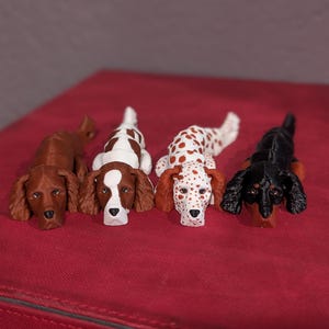 May include: Four dog figurines in various breeds and colors are arranged on a red surface. The dogs include a brown setter, a white and brown setter, a white and red spotted dog, and a black and brown dachshund.
