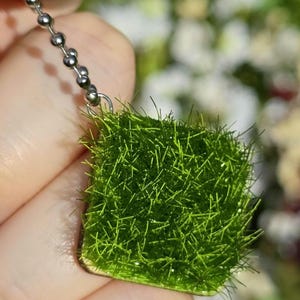 以下が含まれることがあります： 鮮やかな緑色の草のような質感の正方形ペンダント。ペンダントはシルバーのボールチェーンに付いています。ペンダントは手で持たれており、背景は白と緑でぼやけています。