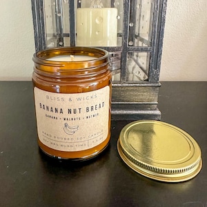 May include: A brown glass jar candle with a gold lid. The label reads "Bliss & Wicks Banana Nut Bread Banana Walnuts Nutmeg Hand Poured Soy Candle 40+ Hrs Burn Time 7.5 oz".