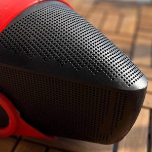 May include: Close-up of a red and black object with a perforated surface. The object has a rounded shape with a black, grid-like pattern on the front. The red portion appears to be a strap or handle.