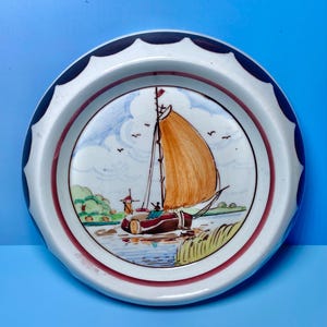 May include: A round ceramic plate with a nautical scene. The plate features a sailboat with an orange sail, a windmill, and a river landscape. The rim is white with a dark blue scalloped edge and a red inner border.