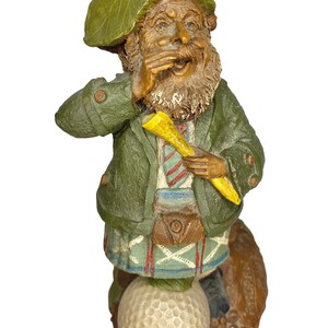 May include: A figurine of a bearded person wearing a green hat, a green jacket, and a plaid kilt. The figure holds a yellow golf tee in one hand and stands with one foot resting on a white golf ball. The figure has a brown beard and moustache.