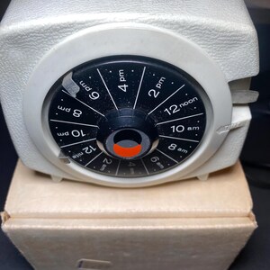 May include: A vintage white and black mechanical timer with a circular dial displaying times from 2 am to 10 pm. The timer sits on a cardboard box. The dial has a black background with white numbers and markings.