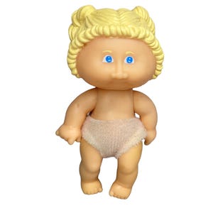 May include: A small doll with light skin, bright blue eyes, and molded yellow hair styled in two pigtails. The doll has a round face and is wearing a simple, light-colored fabric diaper. The figure is depicted in a standing position.