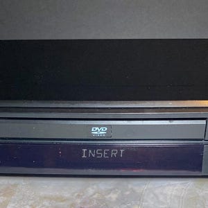 May include: A black Toshiba DVD video player. The front panel displays "INSERT" on a blue screen. Features include a disc tray, control buttons, and the DVD logo. The top is a glossy black finish.