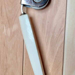 Op de afbeelding: Vintage verchroomde blikopener met een witte handgreep. De blikopener heeft een draaimechanisme en een snijwiel. De handgreep is schuin geplaatst voor een goede grip en hefboomwerking.