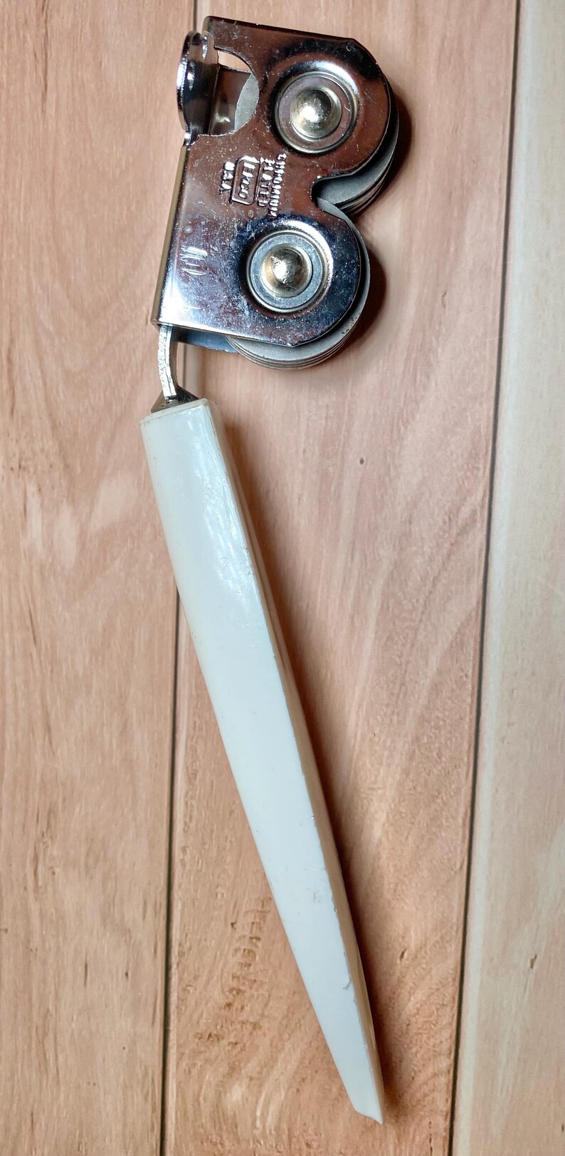 Op de afbeelding: Vintage verchroomde blikopener met een witte handgreep. De blikopener heeft een draaimechanisme en een snijwiel. De handgreep is schuin geplaatst voor een goede grip en hefboomwerking.