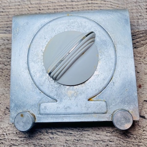 May include: A vintage, square metal object with a circular, gray knob. The knob has ridged detailing and a silver accent. Two small, circular rivets are on the bottom corners. The metal has a worn, aged appearance.