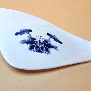 May include: White ceramic rice paddle with a blue floral design. The paddle features a long, curved handle and a wide, flat head. The blue floral design is centered on the head of the paddle and includes a lotus flower and leaves.