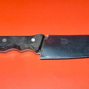 May include: A stainless steel chef's knife with a black handle. The blade is wide and slightly curved, with a sharp edge. The handle is riveted and has a comfortable grip. The knife is set against a bright orange background.