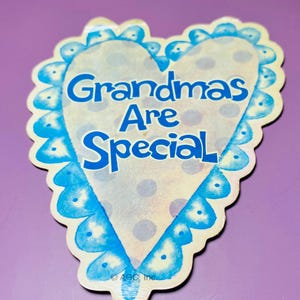 May include: A heart-shaped sticker with the words "Grandmas Are Special" in blue. The sticker has a scalloped edge in light blue and a cream-colored center with blue polka dots. The sticker is on a purple background.