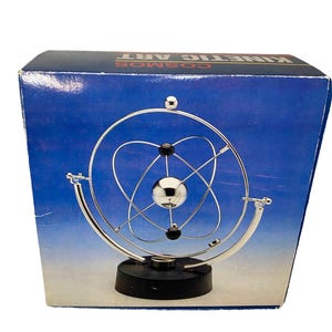 Puede incluir: Una escultura de arte cinético con una esfera central plateada rodeada de anillos plateados giratorios y esferas negras más pequeñas, montada sobre una base circular negra. El producto se muestra frente a una caja azul con el texto KINETIC ART COSMOS.