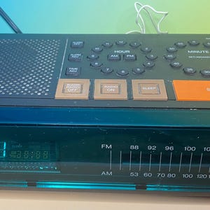 May include: Black Toshiba AM/FM clock radio with a digital display showing the time. The radio has buttons for hour, minute, radio on/off, and snooze. The display shows the time as 12:01.