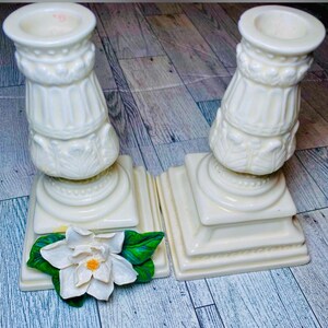 May include: Two white ceramic candle holders with ornate designs. The holders have square bases and are decorated with raised floral patterns. A white magnolia flower with green leaves is placed in front of the holders.