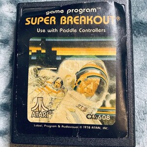 May include: A black Atari game cartridge with a colourful label featuring the text "game program™ SUPER BREAKOUT® Use with Paddle Controllers" and a graphic of two astronauts in space. The cartridge number CX2608 is printed on the label. The label also includes the text "Label, Program & Audiovisual © 1978 ATARI, INC."