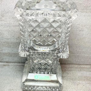 May include: A clear, cut-glass decorative box with a square base and a diamond pattern. The box has a lid and a detailed, geometric design. The glass is transparent, allowing light to pass through, and the box sits on a square base.