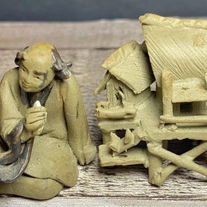 May include: Two small, beige-colored sculptures. One depicts a seated figure holding an object, the other a detailed miniature structure with a roof and chair. The sculptures are on a wooden surface.