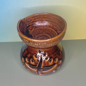 May include: A brown ceramic decorative bowl with the word "Residence" inscribed on the side. The bowl has a rounded top and a base with cut-out designs. The bowl is a dark brown color.