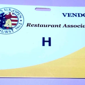 May include: A white and yellow vendor badge from the 2005 U.S. Open at Pinehurst No. 2. The badge features the tournament logo, the text "VENDOR," "Restaurant Associates," and the letter "H."