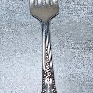 May include: Silverware fork with four tines. The handle features an ornate, floral design. The fork is made of a shiny, silver-colored metal. The background is a textured, light gray surface.