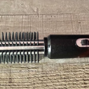 May include: A black and silver hair styling brush with the text "SUPERMAX" printed on the handle. The brush has a cylindrical design with bristles and a power switch with an illuminated light.