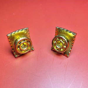 May include: A pair of gold-toned cufflinks. Each cufflink features a square frame with a textured border, surrounding a circular, detailed center. The cufflinks are displayed against a red background.