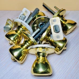 May include: A collection of vintage brass door knobs and hardware. The knobs have a shiny gold finish, with some showing signs of wear. Includes various components like spindles and backplates, suggesting a set of antique door hardware.