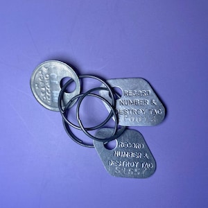 Puede incluir: Un conjunto de etiquetas y llaveros de metal plateado sobre un fondo morado. Las etiquetas tienen el texto "RECORD NUMBER & DESTROY TAG" junto con números. Una etiqueta es redonda y las otras son rectangulares.