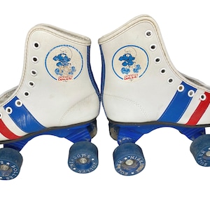 May include: A pair of white roller skates featuring blue and red stripes along the sides and a circular Smurf character graphic on the ankle area. The skates have blue plastic chassis and blue wheels with the text Roller Smurf printed on them.