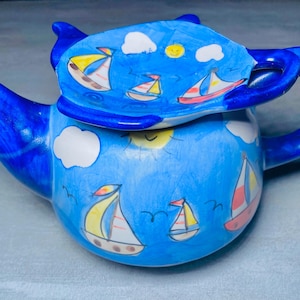 May include: A blue ceramic teapot with a lid featuring a whimsical design of sailboats, clouds, and a sun. The teapot is decorated with hand-painted details in yellow, red, and white.
