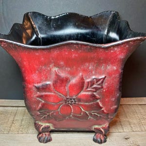 May include: A red decorative planter with a poinsettia design. The planter has a black interior and rests on four small, decorative feet. The planter is approximately 8 inches tall and 8 inches wide.