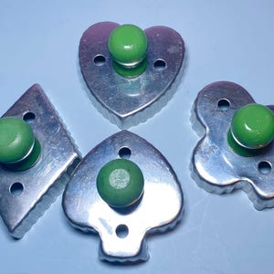 May include: Four metal cookie cutters in diamond, heart, spade, and flower shapes, each with a green wooden knob. The cutters are made of shiny, silver-colored metal and sit on a light blue surface.