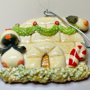 May include: A Christmas ornament featuring a penguin in a white igloo with a green garland and red berries. A red and white candy cane is on the right. The ornament has a metal hook for hanging.