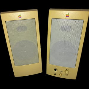 May include: A pair of vintage beige computer speakers, each with a perforated metal grill and a multi-coloured apple logo. One speaker features a headphone jack and volume control. Set against a black background.