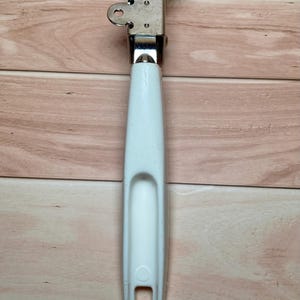 Op de afbeelding: Een vintage blikopener met een witte handgreep en een zilveren metalen mechanisme. De handgreep heeft een gebogen inkeping en een rechthoekige opening aan de onderkant. Het metalen deel heeft twee cirkelvormige openingen.