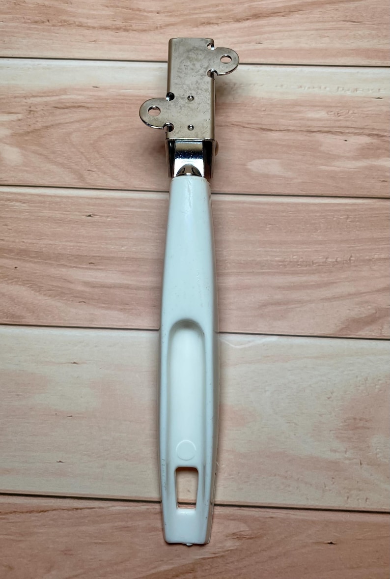 Op de afbeelding: Een vintage blikopener met een witte handgreep en een zilveren metalen mechanisme. De handgreep heeft een gebogen inkeping en een rechthoekige opening aan de onderkant. Het metalen deel heeft twee cirkelvormige openingen.
