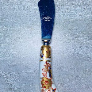 May include: A vintage butter knife with a curved, dark blue blade. The handle is white porcelain, adorned with gold floral patterns and accents of red and blue. The blade is marked "Royal Crown Derby England".