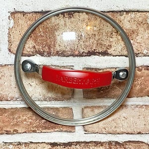 May include: A clear glass Farberware lid with a stainless steel rim and a red plastic handle. The handle is curved and has the Farberware logo in white. The lid is round and appears to be for a pot or pan.