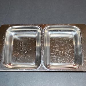 May include: A rectangular, divided serving dish made of stainless steel. The dish has two square compartments and decorative handles with an embossed floral design on each end. The surface shows minor scratches.
