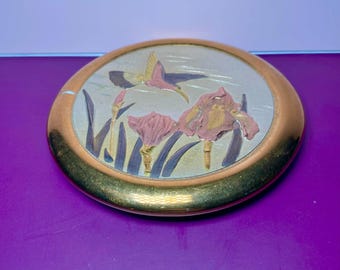 Könnte beinhalten: Eine runde, dekorative Metallplatte mit goldfarbenem Rand. Die Mitte zeigt einen fliegenden Vogel über rosa Irisblüten und grünen Blättern auf silbernem Hintergrund. Die Platte hat einen violetten Hintergrund.