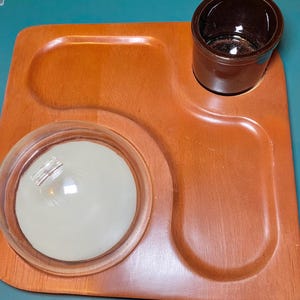 May include: A vintage wooden serving tray with a warm brown finish. It features a recessed area for a small brown ceramic cup and a clear glass bowl with a white, round object inside. The tray has a unique, curved design.