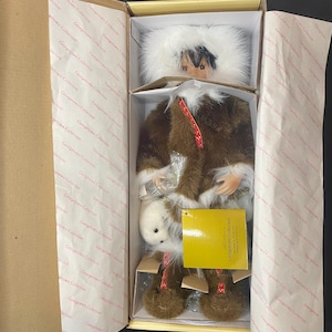 Könnte beinhalten: Eine Puppe aus der Georgetown Collection, verpackt in einer Box. Die Puppe trägt einen braunen, pelzbesetzten Parka mit einer weißen Pelzhaube. Sie hält ein kleines weißes Plüschtier. Die Box trägt das Logo der Georgetown Collection.