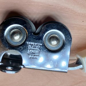 Op de afbeelding: Een verchroomde messenslijper met twee slijpschijven en een cr&egrave;mekleurige handgreep. De slijper heeft de tekst "CHROMIUM PLATED" en "Ekco USA" op het metaal gestempeld. De slijper is ontworpen om messen te slijpen.