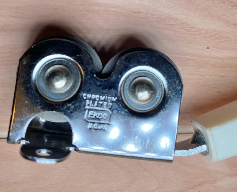 Op de afbeelding: Een verchroomde messenslijper met twee slijpschijven en een cr&egrave;mekleurige handgreep. De slijper heeft de tekst "CHROMIUM PLATED" en "Ekco USA" op het metaal gestempeld. De slijper is ontworpen om messen te slijpen.