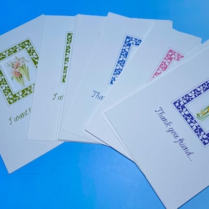 Könnte beinhalten: Eine Sammlung weißer Grußkarten mit floralen Designs und handgeschriebenen Nachrichten. Jede Karte hat einen unterschiedlich farbigen Rand, darunter Grün, Blau und Rosa, und eine Blumenillustration. Die Karten sind auf einer blauen Oberfläche angeordnet.